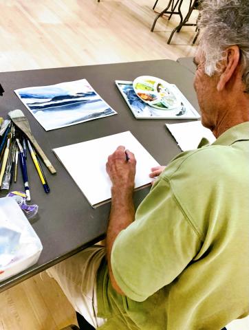 A man sits at a table sketching on a blank sheet of paper, surrounded by watercolor supplies and brushes.
