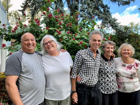 Five smiling adults stand close together in front of a lush flowering bush, enjoying a sunny day.